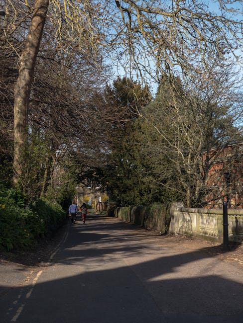 A quiet residential street scene during sunset, featuring a gentle uphill road with a large white multi-story house on the left, displaying multiple windows, a small balcony, and a decorative entrance