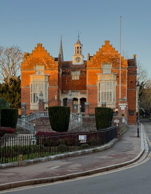 A three-storey historic brick building with decorative gables and large bay windows, situated behind a black wrought iron fence and landscaped with trimmed shrubs and garden beds. The building feature
