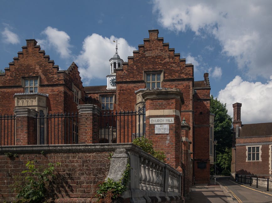 A three-storey historic brick building with decorative gables and large bay windows, situated behind a black wrought iron fence and landscaped with trimmed shrubs and garden beds. The building feature