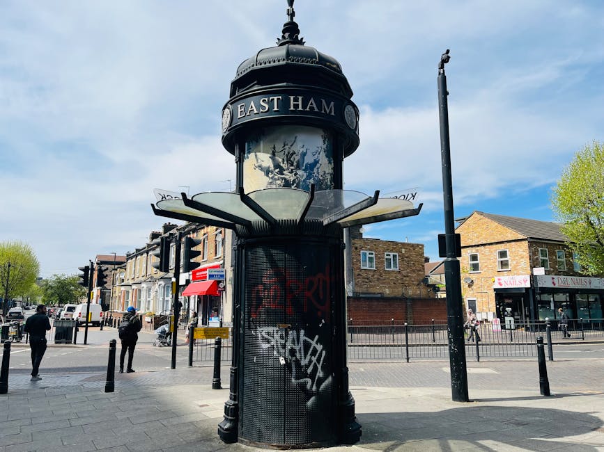 A cylindrical street clock with a black metal base and a decorative top, displaying the text 'EAST HAM' on an oval sign at the top, situated on a paved sidewalk near a busy street intersection in Sout