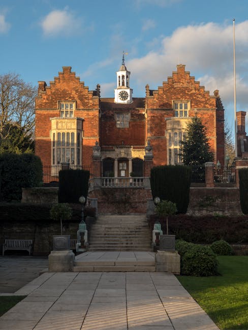 A large, historic brick building with a central clock tower and a decorative gable roof, set behind a stone staircase with metal railings leading up from a paved courtyard. The building is surrounded 