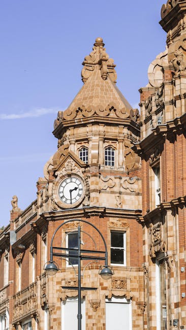 A large, historic brick building with a central clock tower and a decorative gable roof, set behind a stone staircase with metal railings leading up from a paved courtyard. The building is surrounded 